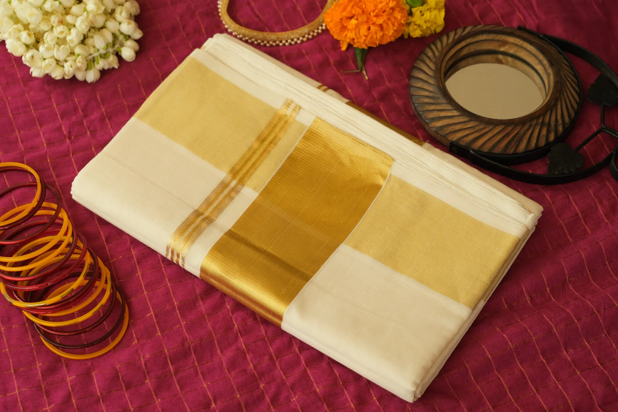 The Kasavu saree folded on a deep red cloth, its golden border beside glass bangles and jasmine