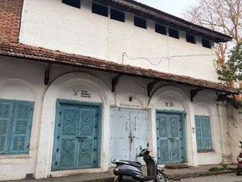A white building with a tiled roof features a row of arched openings along its front. Two of the openings contain ornate blue double doors, while a third holds a plain light blue door. Blue windows with decorative panels flank the doors on either side. Above the central door is a small sign, and above the doors on either side are additional signs with symbols. A scooter is parked on the street in front of the building, and part of another vehicle is visible at the edge of the frame. Bare tree branches appear in the upper right corner against the sky.