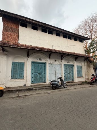 A two-story building with a white facade and a tiled roof stands on a paved street. The upper level features a row of small, dark rectangular openings beneath the eaves, with a section of exposed brickwork visible on the left corner. The ground floor has a covered walkway supported by pillars, housing several arched entrances. Some of these entrances have ornate blue wooden doors with circular patterns, while others are white doors or windows with matching blue shutters. A sign is mounted above one of the central doorways. Parked along the curb in front of the building are two scooters, one grey and one red, with the front wheel of a yellow vehicle visible on the far left edge. Bare tree branches rise against an overcast sky on the right side of the frame.