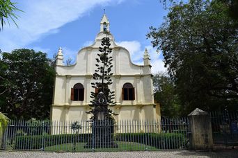 The church from the road: a pale gabled front with scrolled sides and two arched windows with wooden shutters, behind iron railings and a tall conifer.