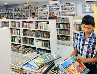 Inside a busy library: full bookshelves down every wall, a boy in school uniform sorting through a stack of paperbacks in the foreground.
