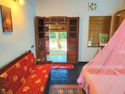 A room with a red floral-upholstered sofa against a pale blue wall, a pink mosquito net over a bed on the right, and open wooden double doors onto a green garden.