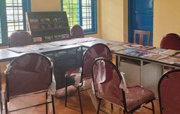 A reading room at KJ Herschel Library with red chairs still wrapped in plastic, tables spread with newspapers and magazines, a magazine rack against the window.