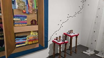 A gallery corner: a painted wooden cupboard stacked with folded, brightly patterned cloth, a line of black ants painted up the wall beside it, and two carved wooden stools holding small tins.