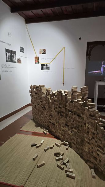 A low wall of small wooden blocks built across a woven mat, some blocks fallen loose in front, with photographs and Bengali text on the wall behind.