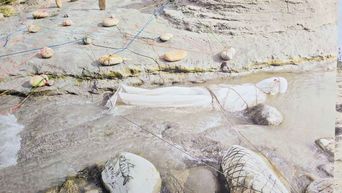 A pale painting of a shallow stony river, coloured threads strung across the rocks and a white cloth net stretched over the water.