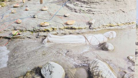 A body wrapped in white cloth lies partially submerged in shallow water at the edge of a rocky bank, surrounded by stones connected by colored strings and wires, with a patterned stone visible in the foreground.