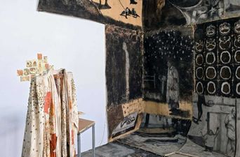 A room hung with huge charcoal-dark cloth panels of figures and rows of dark shapes, with a pale printed robe draped over a stand in the lit corner.