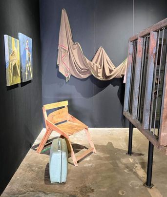 A gallery corner painted dark grey: a fringed brown cloth strung across the wall like a sagging roof, a rough pink-and-yellow wooden chair below it, and two small paintings on the left wall.