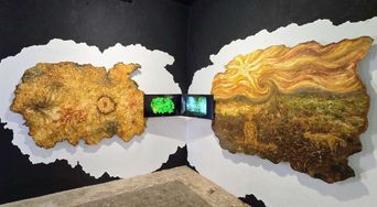 Two large irregular panels of thick ochre and gold textured material mounted on black gallery walls, meeting at a corner where a small video screen plays.