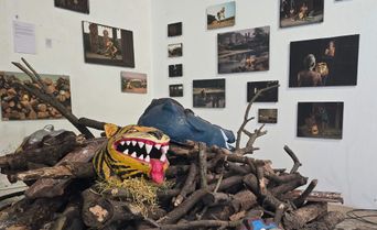 A painted tiger mask with bared teeth and a blue animal mask resting on a heap of cut branches and logs, in a white room hung with framed photographs.