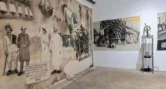 A gallery hung with large charcoal drawings on cloth showing mill workers standing beside cotton-ginning machinery, with a handwritten panel of Hindi text at the lower left and a tall metal machine on the floor to the right.