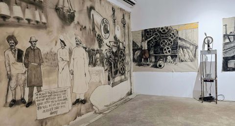 A large mural covers a wall in an indoor space, showing several figures in historical clothing standing beside industrial machinery and hanging objects. To the right, a separate framed artwork depicts a person working among large wheels and spools. A metal stand holding a small device sits on the floor nearby.