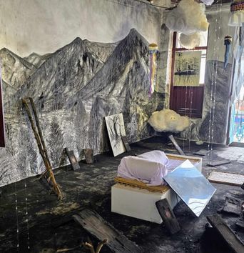 An installation in a bare room: charcoal mountains drawn across the walls, a floor blackened and strewn with burnt timber, stuffed white clouds hanging on strings, and a pale bundle laid on a low plinth.