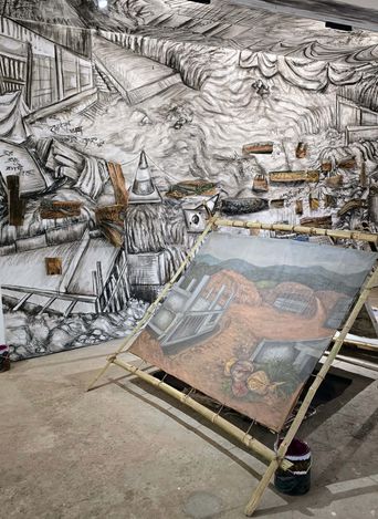 A large, detailed black-and-white mural covers an entire wall, depicting a chaotic scene of collapsed buildings, debris, and draped fabric. In front of the mural, a painting on canvas rests on a bamboo easel, showing a landscape with hills, structures, and figures. A bucket sits on the floor beside the easel.