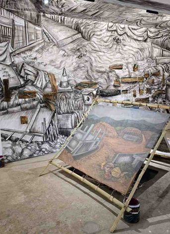 A gallery wall covered by a huge charcoal drawing of collapsed houses and debris, with a painted canvas of a hillside settlement propped on a bamboo frame in front of it.