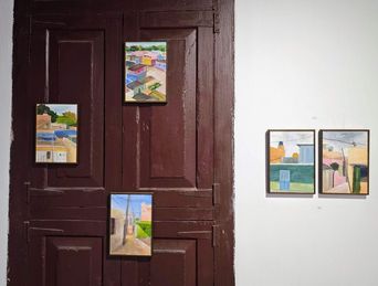 Five small paintings are displayed in a room, three mounted on a dark brown wooden door and two hung side by side on a white wall. Each painting depicts an outdoor scene with buildings, streets, and sky, rendered in a colorful, stylized manner.