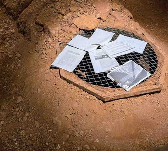 Printed pages and a plastic folder lying scattered on a wire mesh grille set into bare red earth, lit from one side.