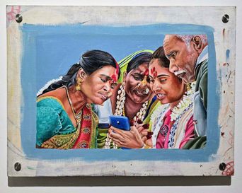 A painting on a rough wooden board: four people in saris and garlands crowding together to look at a blue mobile phone held in one of their hands.