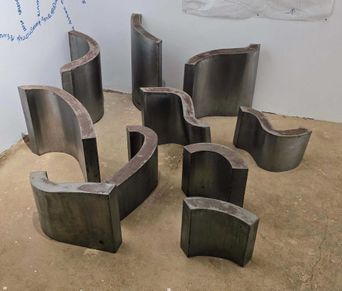 Eight dark curved metal sculptures, each like a section cut from a large drum, standing apart on a bare earth floor in a white room.