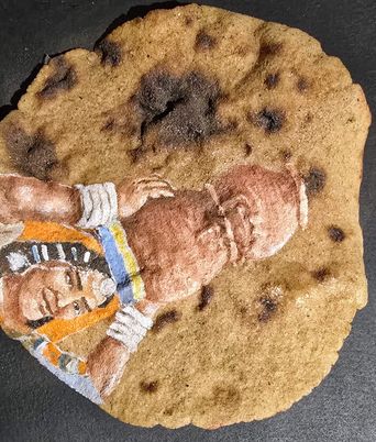 A round, flat baked good with dark spots across its surface, featuring a painted image of a person in profile wearing a striped head covering and a colorful garment, lying on their side with one arm bent and resting near their head.