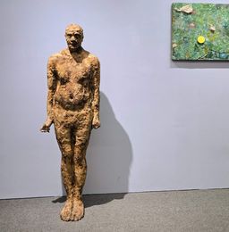 A rough-textured bronze standing figure sculpture at St. Andrew’s Parish Hall, a small green abstract painting on the wall behind it.