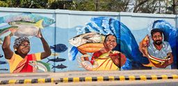 A street wall mural showing fisherwomen and a fisherman carrying large fish, with a blue ocean wave painted behind them, part of the Guardians of Fort Kochi mural.