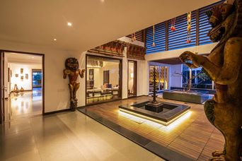 An open-plan hall with a sunken water feature and lit wooden deck, a large carved wooden figure at right, a carved wooden lion-like statue beside a doorway, and louvred windows above.