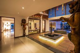 An open-plan hall with a sunken water feature and lit wooden deck, a large carved wooden figure at right, a carved wooden lion-like statue beside a doorway, and louvred windows above.