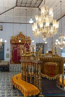 The synagogue’s raised reading platform draped in gold cloth, seen beneath a large crystal chandelier, with the gilded torah ark against the far wall.
