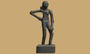 A small dark bronze figurine of a standing nude woman with a segmented, coiled left arm, photographed against a plain tan background.