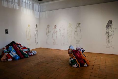 A gallery space with white walls and a tiled floor features faint pencil sketches of human figures along the back wall. On the floor, two large piles of colorful folded fabrics sit in opposite corners, one near the left wall and one closer to the center-right. A small dark sign is mounted on the left wall near the floor.