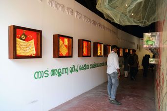 A visitor looks along a gallery corridor at Aspinwall House lined with small illuminated framed artworks, Malayalam text stencilled on the wall above.