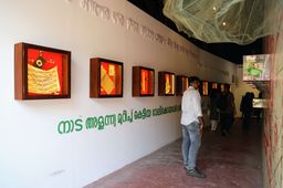 A visitor looks along a gallery corridor at Aspinwall House lined with small illuminated framed artworks, Malayalam text stencilled on the wall above.