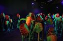 A dark gallery room at Aspinwall House filled with tall sculptural plant-like forms in glowing orange and green under UV light.