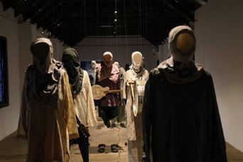 An installation of headless figures in layered pale robes and headscarves standing in rows down a gallery, one holding a guitar, sandals on the floor between them.