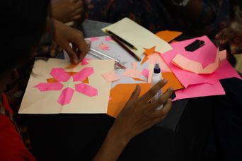 Hands at a table cutting and arranging pink and orange paper shapes into a symmetrical star pattern, with scissors and a glue bottle beside them.