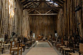 A long, narrow hall with walls and ceiling constructed from vertical bamboo poles tied together, supported by exposed wooden beams overhead. Rows of wooden chairs are arranged on both sides of a central aisle leading toward a raised platform at the far end, where more seating and equipment are visible. Light streams in from openings near the roofline, illuminating the space.