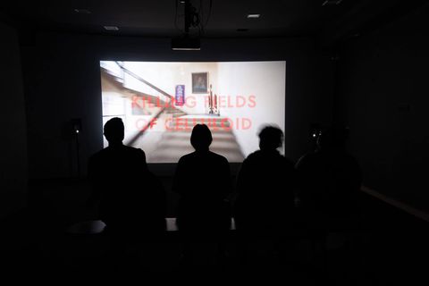 Visitors sit in silhouette watching a video projection titled ‘Killing Fields of Celluloid’ showing a staircase inside a building, at SMS Hall.