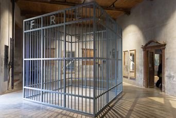 A large steel cage standing alone in a bare room, the word FREE spelled down its bars, throwing striped shadows across the wooden floor.
