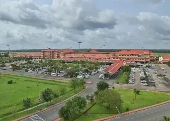 An aerial view of Cochin International Airport’s red-roofed terminal building, with a large car park, access roads and green lawns in the foreground under a partly cloudy sky.