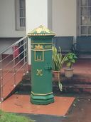 A green-and-gold Victorian-style pillar postbox (anchal box) on a covered porch, with potted plants and a metal railing beside it.