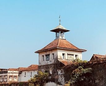 A whitewashed clock tower with a tiled pyramidal roof and weathervane, rising above a stone perimeter wall, part of the Paradesi Synagogue complex in Mattancherry.