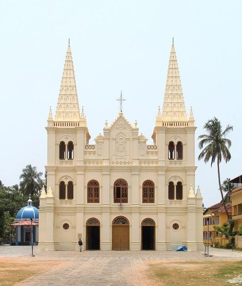 The cream twin-spired facade of Santa Cruz Basilica against a clear sky, with a blue-domed shrine to its left and a coconut palm to its right.