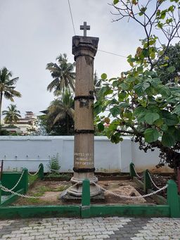 A weathered stone pillar topped with a cross, inscribed ‘Erected by J.E. Winckler, Port Officer’, standing behind a low green fence with palm trees in the background.