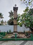 A weathered stone pillar topped with a cross, inscribed ‘Erected by J.E. Winckler, Port Officer’, standing behind a low green fence with palm trees in the background.