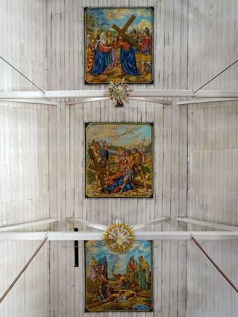 Three framed Stations of the Cross paintings mounted in a row along a white wooden vaulted ceiling.