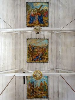 Three framed Stations of the Cross paintings mounted in a row along a white wooden vaulted ceiling.