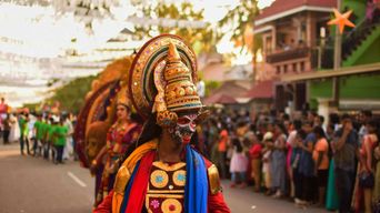 A Kathakali performer in full green and gold headdress and painted face walks down a street in procession while a crowd watches from the kerb.