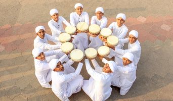 Twelve young men in white kurtas and caps sit in a ring on paving, seen from above, each holding a duff, a round frame drum, towards the centre.